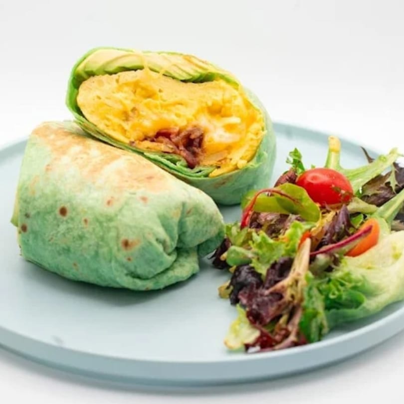 Green tortilla breakfast burrito with scrambled eggs, bacon, avocado, and a side salad.