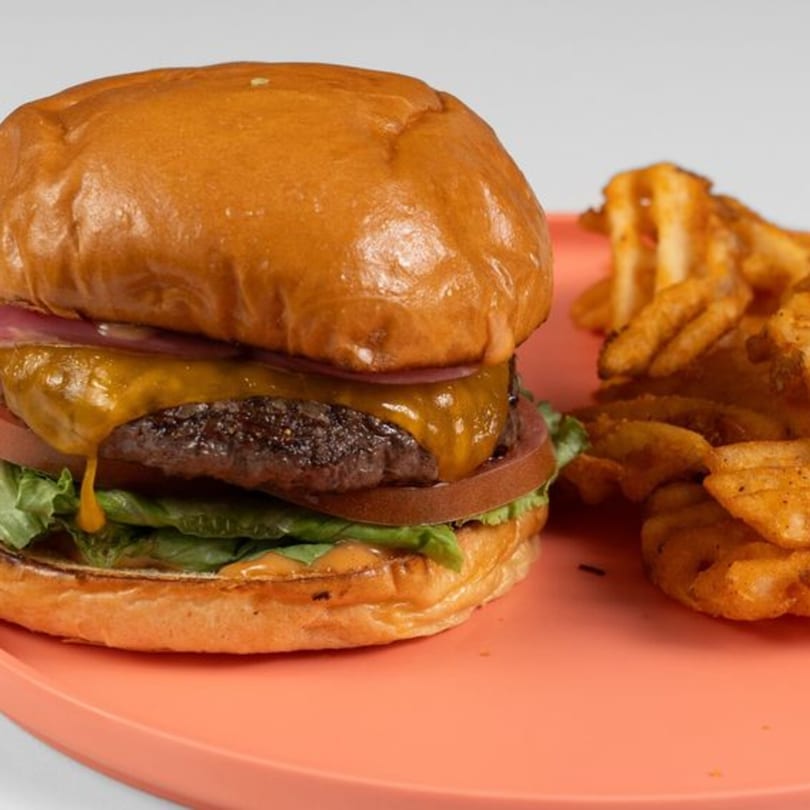 Cheeseburger with melted cheese, lettuce, tomato, and red onion, served with waffle fries on a coral plate.