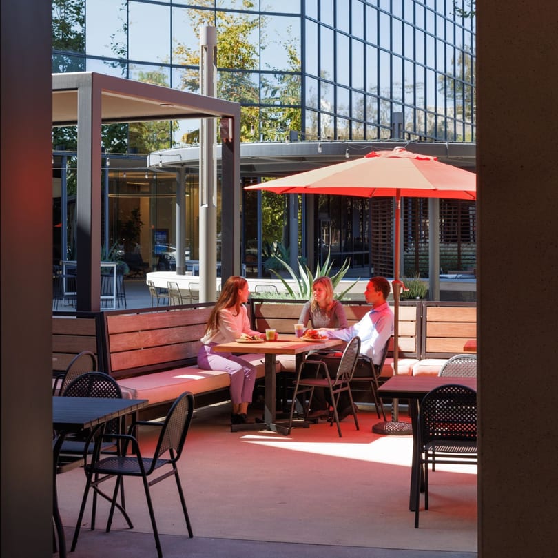Three people enjoying lunch at an outdoor patio table with a red umbrella and modern architecture.