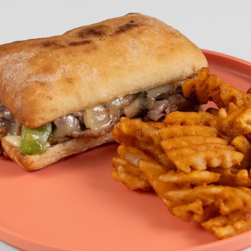 Cheesesteak sandwich with green peppers and onions, served with waffle fries on a coral plate.